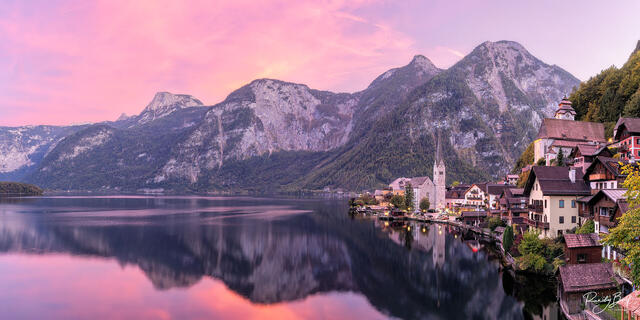 panoramic photograph of Hallstat, Austria at sunset with pink and purple clouds reflecting in the lake.