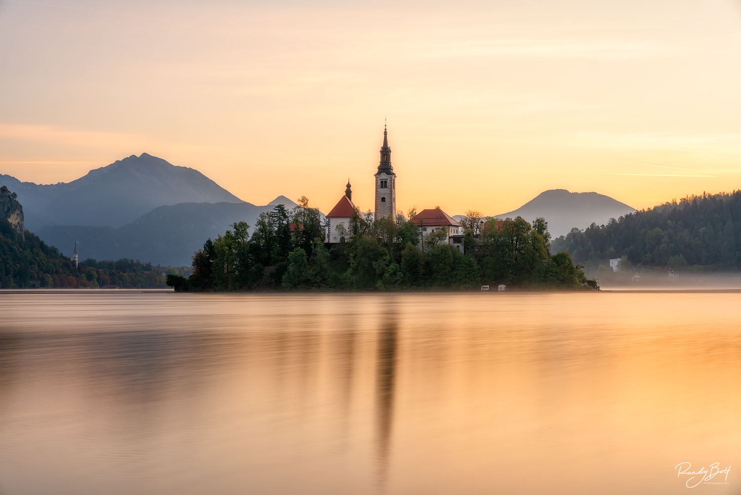 sunrise over the Lake Bled Monastery with mist and colorful hues in the sky.