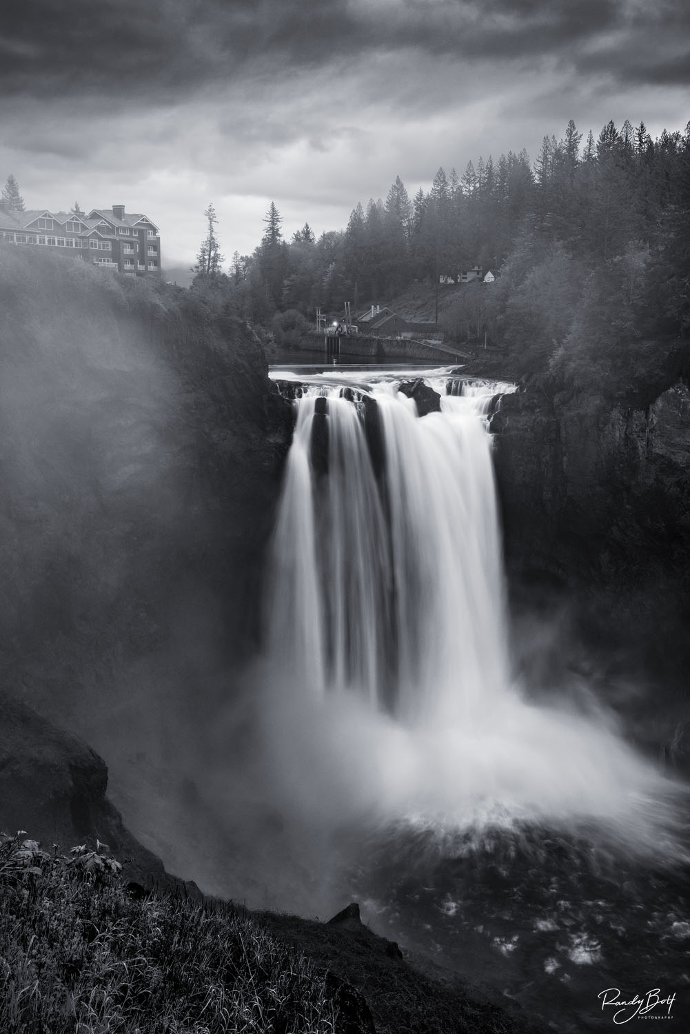 Snoqualmie Falls Storm fine art photograph of Snoqualmie Falls, captured in black and white. The waterfall is seen from a distance, cascading down a rocky cliff into a pool of water.